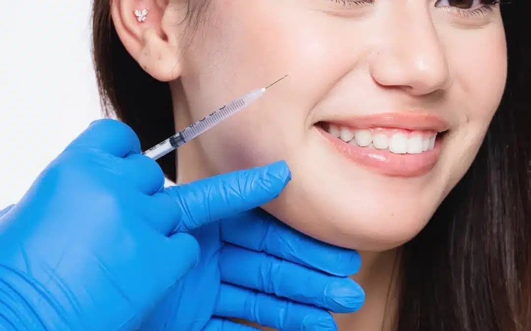 Smiling woman receiving a cheek injection from a practitioner wearing blue medical gloves against a white background.